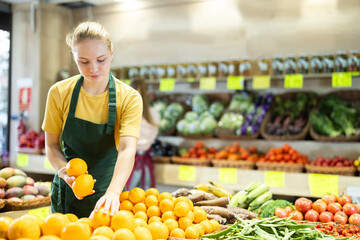 Teenage girl seller in apron puts fresh oranges on display in vegetable shop