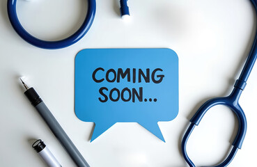 Blue speech bubble with text COMING SOON sits on white table next to medical stethoscope and pen. Announcement of new medical innovation or healthcare service.