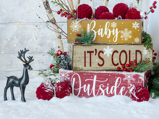 Christmas stacked crates on snow, holiday text Baby It’s Cold Outside, black deer and velvet burgundy ornaments, birch tree, festive fairy lights, greenery, red berries on white plank rustic backdrop