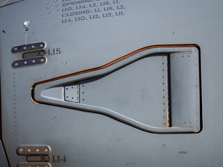 Close-up of metallic aircraft fuselage surface with contoured access panel, fasteners, and hinge mechanisms visible in detail