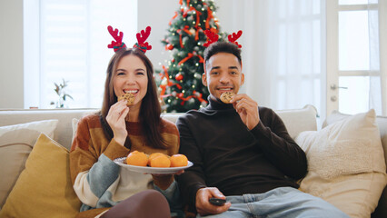 Multiracial couple shares cookies and pastries while sitting on a couch and watching TV, celebrating the festive season next to a beautifully decorated Christmas tree.
