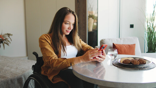 A young woman in a wheelchair happily uses her smartphone while sitting at a white table with cookies and a glass of water in a well-lit space.
