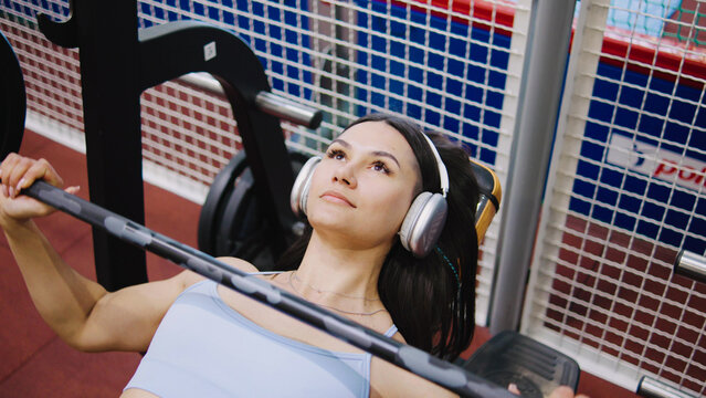 A woman exercises on a bench press in a gym. She is wearing headphones and concentrating on her workout while surrounded by gym equipment during the day.
