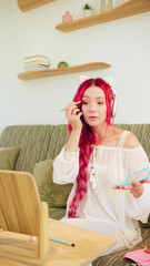 Pink-haired woman focuses on her makeup application, seated on a green couch in a stylish living room, wearing headphones and looking into a mirror.