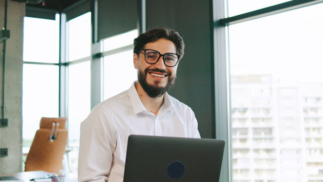 Smiling businessman working on laptop in modern office during daylight with large windows and urban view