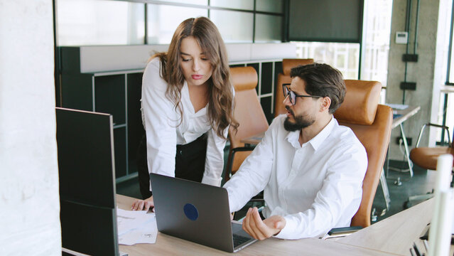 Two colleagues engage in a discussion about a project at a sleek office desk, with a laptop open and paperwork nearby, showing teamwork in a contemporary environment. - Powered by Adobe