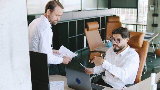 Two business associates engage in a discussion about a project while seated at a desk in an open-plan office space with large windows.