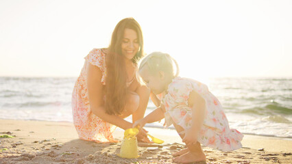 A joyful mother helps her young daughter build a sandcastle on a sunny beach, with ocean waves gently lapping at the shore during late afternoon light.