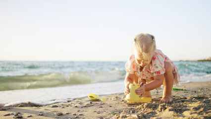 A toddler is engaged in sandy activities at the beach, concentrating intently while constructing a sandcastle with a yellow bucket as waves gently lap nearby.