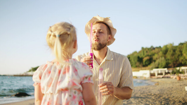 A man engages playfully with his daughter at the beach, blowing bubbles as the sun sets in the background, generating a picturesque and joyful atmosphere. - Powered by Adobe
