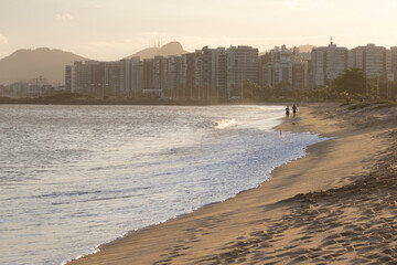 A peaceful golden hour view at Camburi Beach in Vit&oacute;ria, Esp&iacute;rito Santo, Brazil, with two people walking along the sand by the sea and the city skyline in the background
