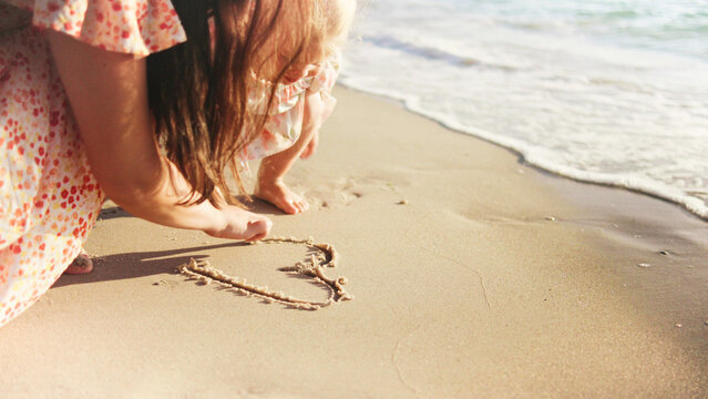 Mother and daughter drawing a heart in the sand by the ocean during a sunny day at the beach