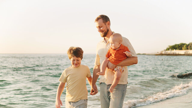 A father walks along the shoreline with his older son while holding a baby. The calm ocean waves lap at their feet as the sun sets in the background.