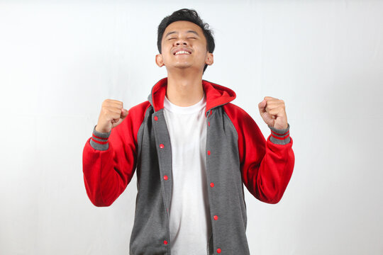 Asian man with a happy expression wearing a red and gray hoodie, celebrates a success with his fists clenched, isolated on a white background.