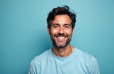 Obraz premium Happy man with dark hair and beard smiles broadly. He wears light blue t-shirt and stands against plain blue background. His expression is friendly and confident, suggesting positivity and success.