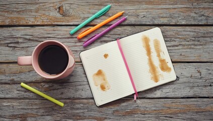 A top-down view of a coffee cup and a notebook with colorful pens, featuring coffee stains on the open pages.