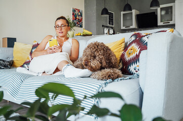 Maltipoo dog lying on sofa while middle aged female in glasses browsing her smartphone relaxing on comfortable living room couch enjoying quiet peaceful moment together in cozy home environment