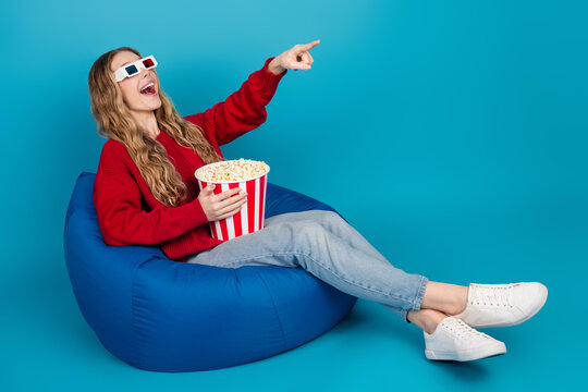 Adorable cheerful young woman with popcorn and 3D glasses watching movies on a bean bag with a positive vibe