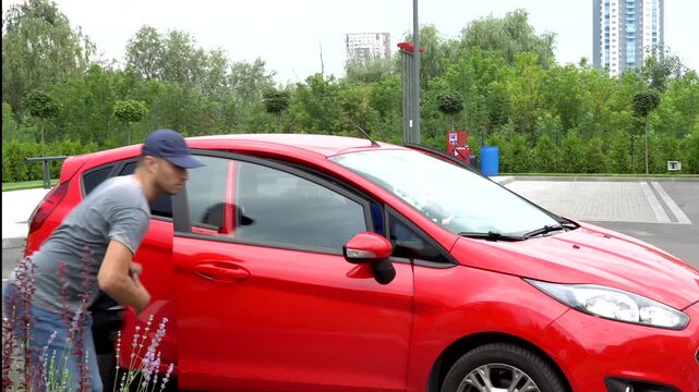 Man in grey clothes and cap comes to car and openes door. He steals purse and run out. Young woman comes out of red car and looks at thief with unhappy. She is upset. Model leans on car.