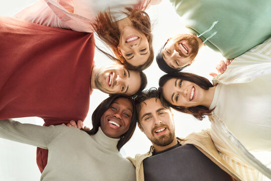 Fototapeta Group of happy diverse friends standing in circle, hugging each other and looking down at camera. Bottom view of smiling multiracial people embracing, showing friendship, unity, cohesion and teamwork.