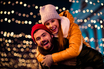 Joyful couple outdoors under festive lights share a playful moment creating a warm Christmas scene...