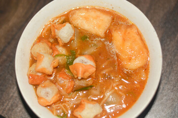 Spicy Indonesian seblak with chicken feet, tofu, sausage, crackers, lime, and vegetables in chili broth. Top view on white background.