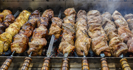 Several rows of skewered meat shashlik or kebabs with wooden handles are grilling over hot coals, releasing smoke in a close-up shot of a food stall