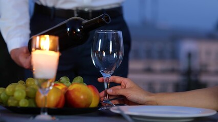Camera moving back from male holding a bottle of wine serving drink to girlfriend at romantic date in evening. Man pouring red wine into glass on roof at date. Love relations