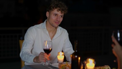 Handsome young male sitting at table with candles at romantic dinner with girlfriends drinking wine and talking. Happy man having romantic date with beloved woman in evening