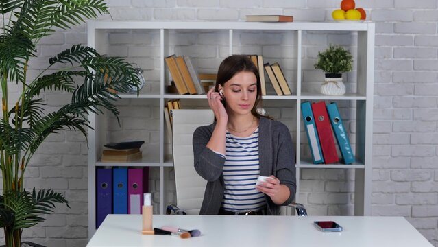 Smiling female blogger shooting beauty vlog on mobile phone, waving hand at camera, greeting subscribers. Woman using wireless headphones
