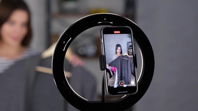 Smiling female vlogger recording video for her subscribers while holding and showing new clothes at home. Woman filming advertisement indoors