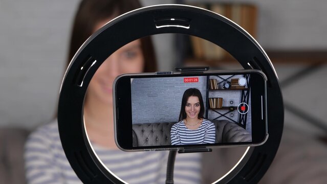 Smiling lady sitting on sofa and talking online looking at phone. Video vlogger recording vlog at home. Lifestyle vlogging concept. Focus on display of phone. video - Powered by Adobe