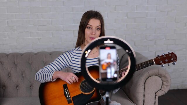 Young female influencer playing guitar during live video streaming for her audience from smartphone at home while sitting on sofa. Art, hobby concept. video - Powered by Adobe