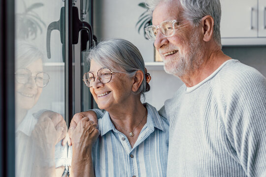 Happy bonding loving middle aged senior retired couple standing near window, looking in distance, recollecting good memories or planning common future, enjoying peaceful moment together at home.