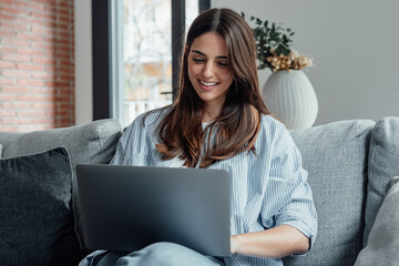 Concentrated millennial girl sit on couch working on laptop browsing internet at home during lazy...