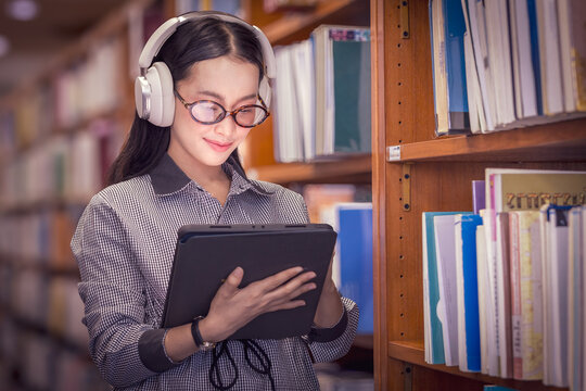 Asian woman with headphone reading book in library for education, studying or research in school, university or college campus. Focus, book and student at bookshelf for learning or knowledge 