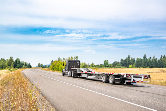 American classic black big rig semi truck tractor with empty step deck semi truck running on the summer winding road