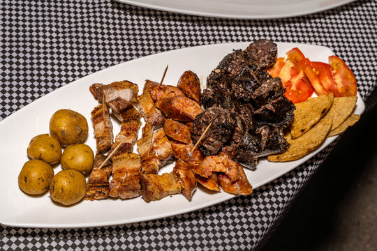 Grilled pork, beef, chorizos and morcillas with potatoes, tomato slices and fried plantains on white plate over checkered tablecloth in Colombian home