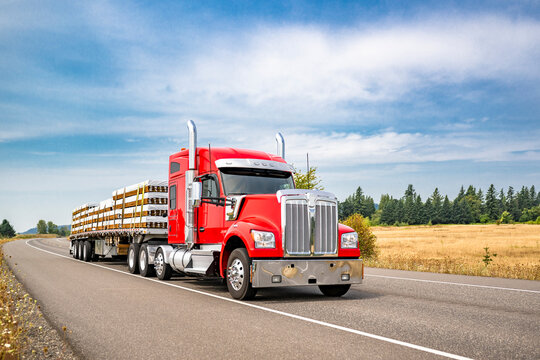 Fototapeta Bright red bonnet big rig semi truck with chrome accessories transporting fastened lumber cargo on flat bed semi trailer running on the narrow summer road