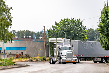 Classic style powerful white big rig semi truck with roof windows and chrome accessories transporting cargo in flat bed conestoga semi trailers turning on the local road in industrial area