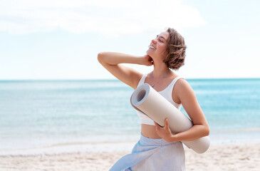 Smiling slim woman in sporty active clothes standing by sea with rolled yoga mat under bright sky. She is enjoying healthy outdoor lifestyle and fresh air near the ocean