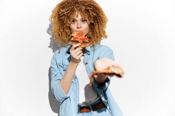 Young woman with curly hair enjoying pizza, wearing denim outfit, demonstrating joy and indulgence, bright white background, food concept