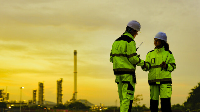 male and female engineer in high-vis gear and hard hats use a tablet. They are collaborating at an industrial factory or refinery site during a golden sunset. - Powered by Adobe