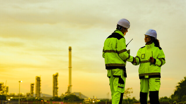 male and female engineer in high-vis gear and hard hats use a tablet. They are collaborating at an industrial factory or refinery site during a golden sunset.