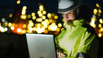 A focused female industrial worker in a hard hat and high-vis jacket uses a laptop at night. She is...