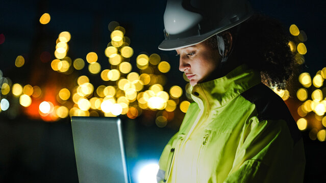 A petrochemical engineer in PPE monitors refinery operations during a night shift. She uses a laptop for real-time data analysis in front of lit-up processing infrastructure. - Powered by Adobe