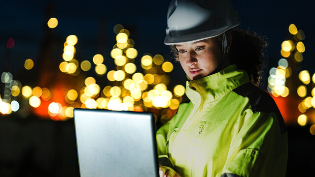 A petrochemical engineer in PPE monitors refinery operations during a night shift. She uses a laptop for real-time data analysis in front of lit-up processing infrastructure.