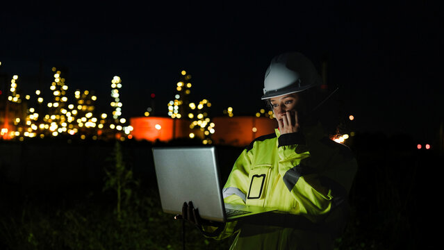 A petrochemical engineer in PPE monitors refinery operations during a night shift. She uses a laptop for real-time data analysis in front of lit-up processing infrastructure. - Powered by Adobe