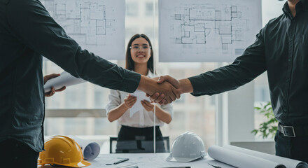 Architects shake hands sealing deal with blueprints and hard hats on table, celebrating successful project milestone