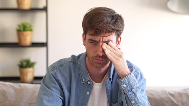 Tired young man feel pain eyestrain holding glasses rubbing dry irritated eyes fatigued from computer work, stressed man suffer from headache bad vision sight problem sit at home table using laptop
